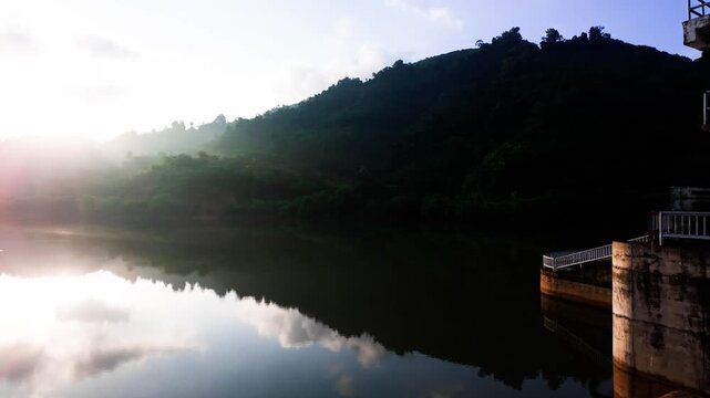 People walk along the path at Da Lay Irrigation Reservoir in Lam Dong, Vietnam. The sun rises over the water, creating reflections on the calm surface. Green hills surround the area.