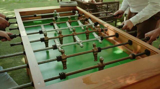 Several pairs of hands working the rods of a wooden matraquilhos table set up on a lawn, the green pitch and metal players in sunlight during a garden party.
