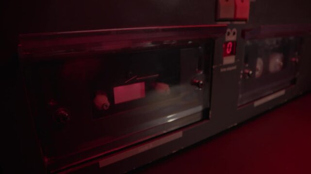 Hand loads a cassette tape into a dual deck player under red light. Gritty retro audio recording scene, ideal for police interrogation, wiretap evidence, crime mystery, and forensic thrillers.