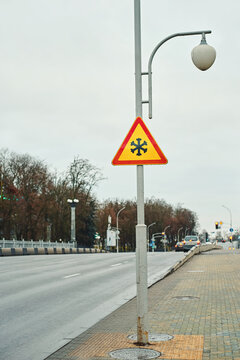 Yellow warning sign with snowflake icon mounted on street lamp pole on urban sidewalk preparing drivers for slippery winter roads. Snowflake traffic sign on pole warn motorists of ice hazards on road