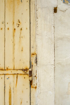 Aged Cream Painted Wooden Door Planks, Rusty Metal Hinge and Flaking Paint Next to Cracked Stucco Architectural Wall