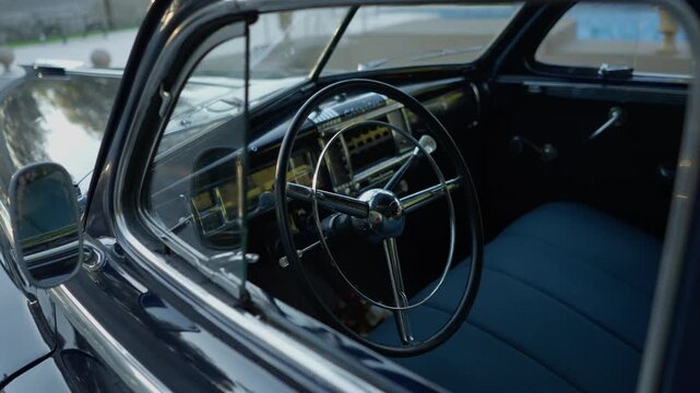 Classic black sedan seen through the open window, with a thin spoked wheel, painted dashboard, bench seat and side mirror.