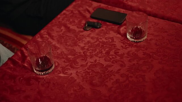 Moroccan salon table with half drunk glasses, a smartphone and a vehicle fob on patterned crimson cloth, a guest's knee at the edge.