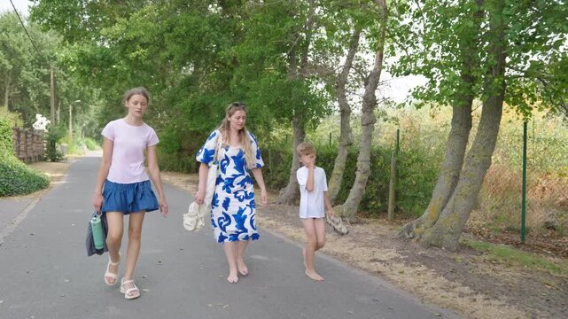 Woman and two children walking along a tree lined path carrying shoes and beach belongings