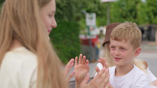 A teenage girl is teaching a boy a hand clapping game slow motion while sitting on a bench in the park with mother