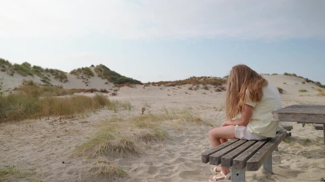 Girl sitting on a wooden bench among coastal sand dunes slow motion adjusting shoes gets up and walks away rear view