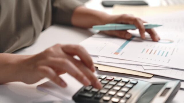 Female accountant checking financial documents while using calculator at office desk, reviewing records, verifying figures and calculating expenses for accounting, finance, tax preparation and bookkee