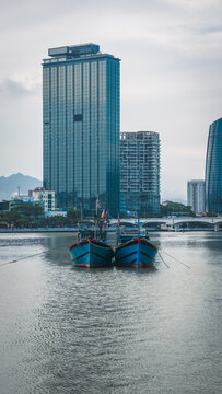 traditional wooden fishing boats on river near modern city skyline, blue vessels moored inside urban bay against towering glass skyscrapers of Danang