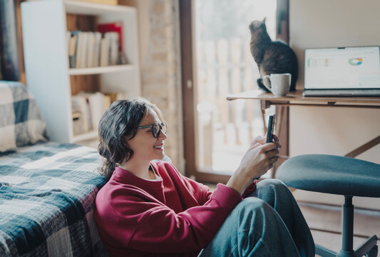 Smiling young woman in glasses looking at smartphone screen with bookshelf behind her