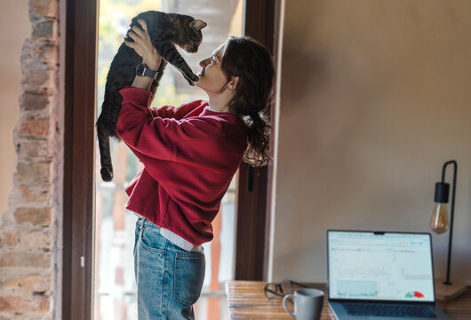 Woman standing by window holding cute tabby cat in front of her face near desk