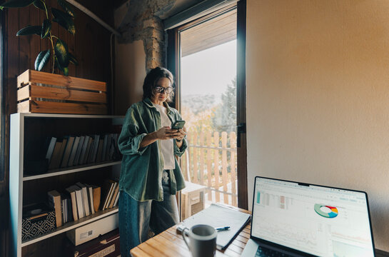 Young woman in green shirt standing by desk with laptop and looking at smartphone screen