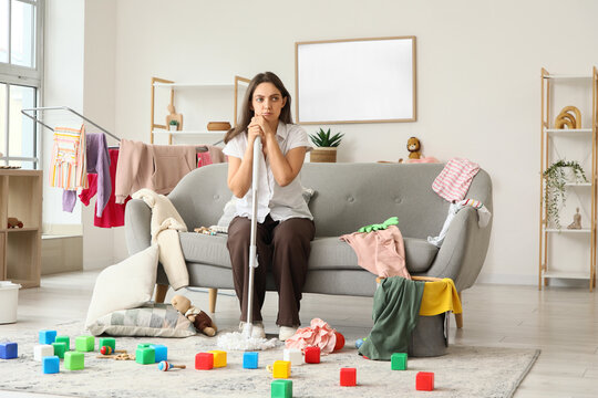 Burnout and domestic fatigue, exhausted woman sitting on sofa with mop in messy living room surrounded by laundry and children toys after housework
