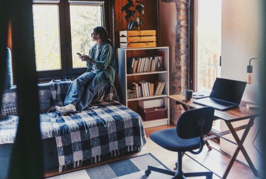 Wide angle view of young woman in green shirt sitting on windowsill with smartphone looking out window