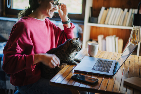 Young woman in glasses and red sweatshirt working from home on laptop with cat on lap