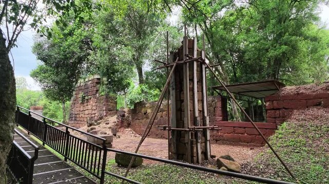 Camera pans left along railed visitor walkway past a sandstone pillar braced with wooden supports for conservation at the Loreto Jesuit ruins with green trees surrounding.