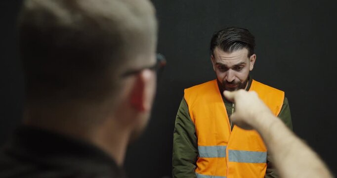 A manager gestures sternly toward an employee wearing an orange safety vest in a dramatized workplace reprimand scene
