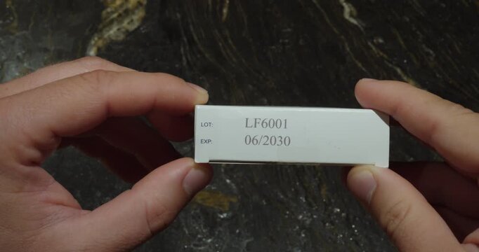 Close up of hands holding a medicine box and inspecting the printed lot number and expiration date on its side
