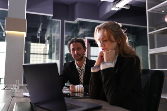 Both colleagues stare flatly at the laptop with heavy eyes, exhaustion settling over the desk as the night stretches on in the office in a bright modern coworking office.