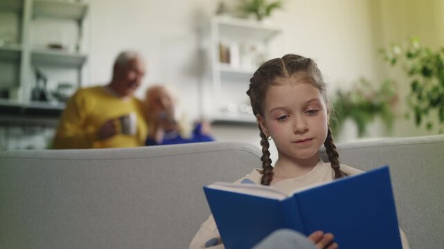 Little girl reads book while grandparents drink tea, educational family time
