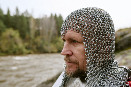 Middle aged CaucLatin male reenactor wearing chainmail armor outdoors near river, portraying medieval knight. Image suitable for history education, gaming and fantasy advertising, close up shot