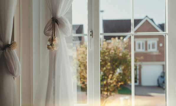 Window autumn view from inside home, white curtains framing a suburban house and golden fall foliage in soft sunlight, peaceful interior background with seasonal scenery, cozy atmosphere and daylight.