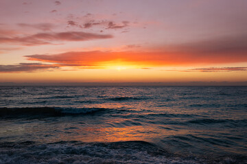 The magnificent colorful sunset sky with vibrant purple, pink, and orange hues reflected in the wet sand and gentle waves of the sea coast © castenoid