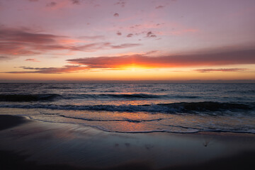 The magnificent colorful sunset sky with vibrant purple, pink, and orange hues reflected in the wet sand and gentle waves of the sea coast © castenoid