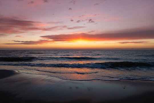 The magnificent colorful sunset sky with vibrant purple, pink, and orange hues reflected in the wet sand and gentle waves of the sea coast