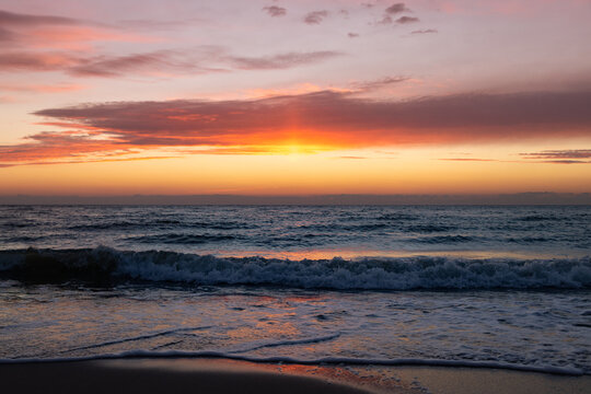 The magnificent colorful sunset sky with vibrant purple, pink, and orange hues reflected in the wet sand and gentle waves of the sea coast