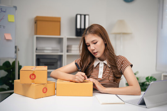 Young woman preparing cardboard box for shipping writing on label with pen in modern home office workspace