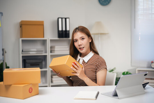 Young woman sitting at desk holding cardboard box preparing home delivery package with laptop and notepad nearby