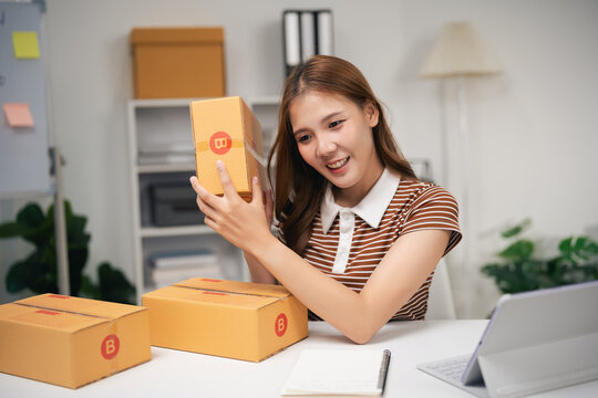 Young woman smiling while examining small parcel box at home workspace with laptop and notebook on white table