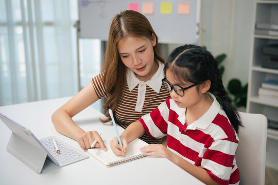 Young woman tutoring school age girl with notebook and tablet in modern bright home study setting
