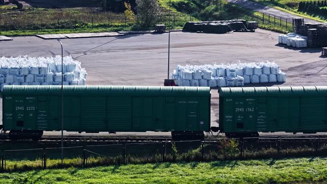 Green freight boxcars standing on railway tracks beside paved storage yard with rows of white bulk bags, fence and green grass on sunny day