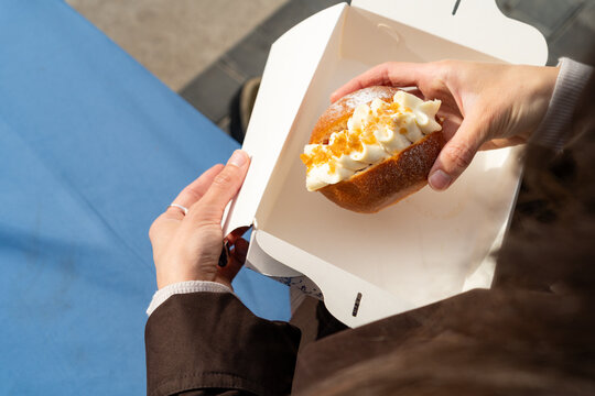 Close-up of a woman sitting outdoors holding an open cardboard takeaway box and a sweet brioche bun filled with whipped cream, caramel drizzle, and a dusting of icing sugar