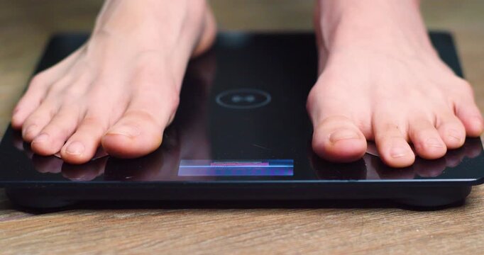 Close-up footage shows a teenager boy stepping onto electronic bathroom scales to check his weight, as part of a personal health and fitness routine at home each morning.