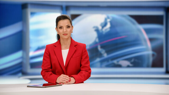 Portrait of a Female News Anchor Presenter Sitting at a Studio Desk With a Tablet and Folded Hands, Delivering World News Headlines to the Camera During a Live Evening Broadcast in a Television Studio