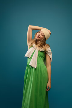 Relaxed blonde woman resting hand on head against teal studio background in green dress. Summer fashion, personal wellbeing, lifestyle editorial concept.