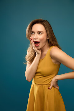 Astonished woman touching cheek with hand on hip against teal studio background. Retail announcements, reaction content, casual fashion concept.
