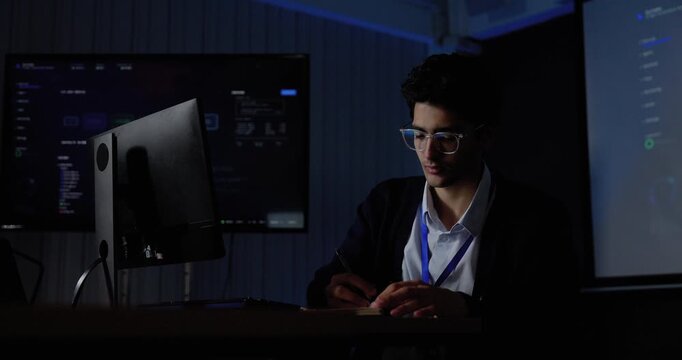 Young Asian man types at computer workstation, leans back and touches head while pausing from software work, conveying office fatigue, workplace stress, system analysis, digital workflow planning.