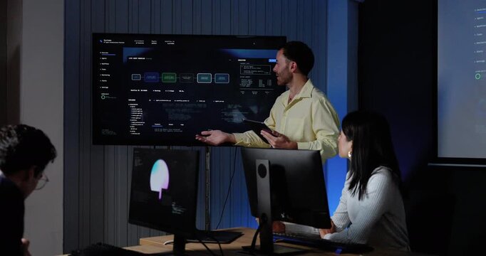 Adult male technology colleagues discuss software dashboard, point toward system interface, and exchange ideas while female colleague works at computer during digital strategy meeting in office.