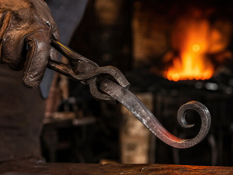 A gloved hand expertly bends a newly forged dark metal hook with tongs over a dark grey anvil within a dimly lit blacksmith&rsquo;s workshop.
