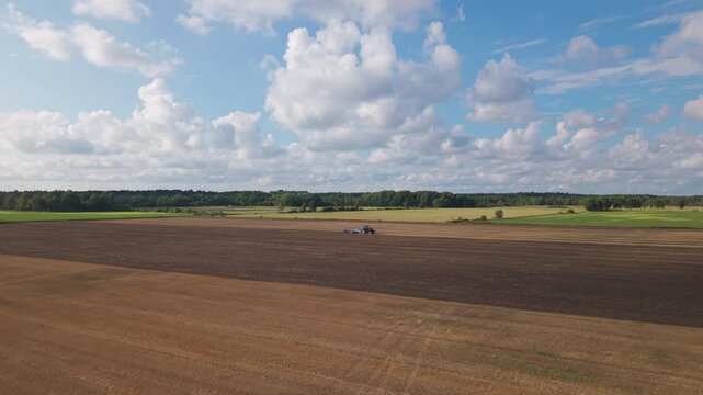 An elevated view shows a tractor with soil preparation equipment amid parallel strips of stubble and worked soil, bordered by green fields and woodland.