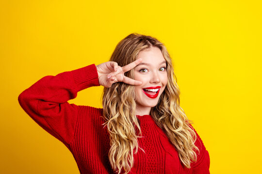 Young woman smiling in a red sweater making a peace sign against a bright yellow background, playful portrait conveying happiness, confidence and fun with blonde hair and bold red lipstick