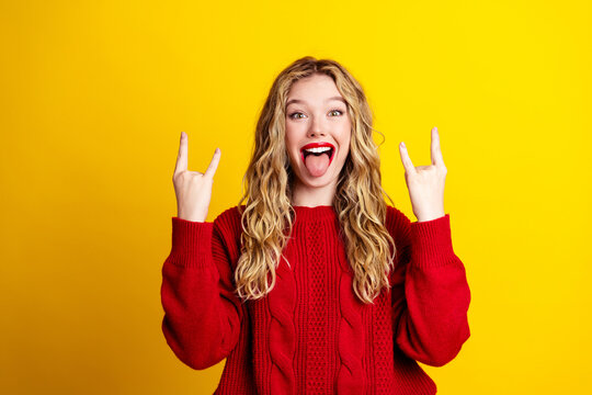 Playful young woman in red sweater making rock-on gesture and sticking out tongue against bright yellow background