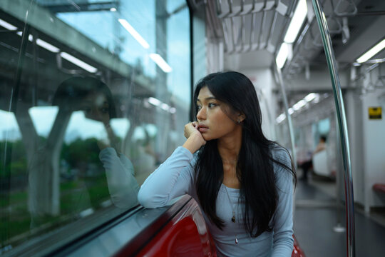 Pensive young Asian woman looking out window while riding urban metro train alone
