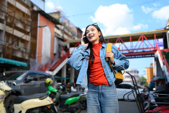 Happy Asian woman traveler talking on phone while exploring colorful urban street in Asia with yellow backpack