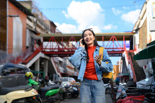 Happy Asian woman traveler talking on phone while exploring colorful urban street in Asia with yellow backpack