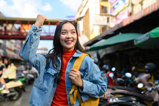 Cheerful Asian female tourist pumping her fist in excitement on a bustling urban street, perfect for travel, tourism, and adventure campaigns.