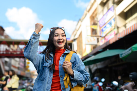 Excited young Asian woman traveler celebrating with fist pump on vibrant city street with backpack